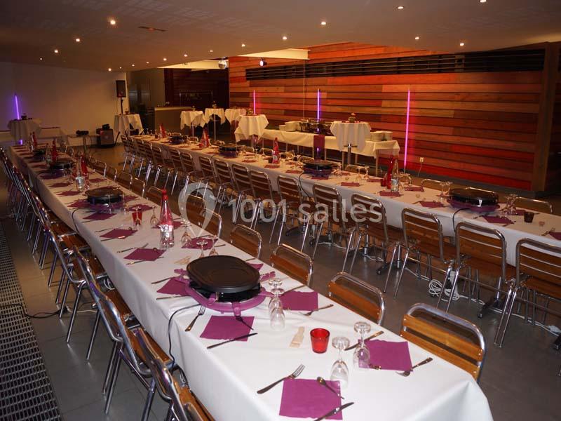 Salle aménagée avec de longues tables dressées, nappes blanches, serviettes violettes et chaises en bois, prête pour un…