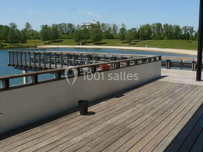 Terrasse en bois avec vue sur un ponton au bord d'un lac, entouré de pelouses et d'arbres sous un ciel dégagé.