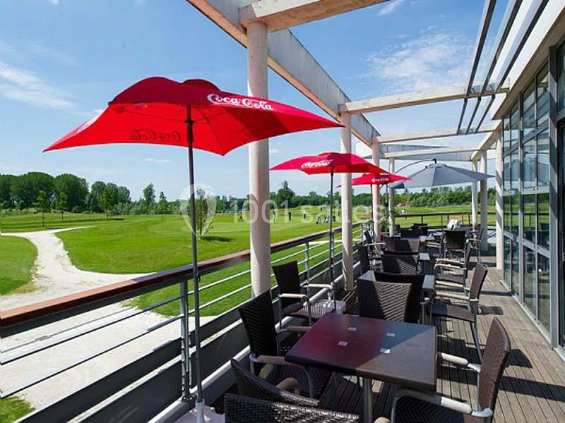 Terrasse avec tables, chaises en osier et parasols rouges, donnant sur un terrain de golf sous un ciel dégagé.