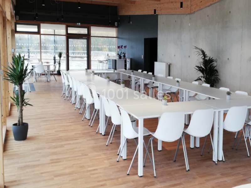 Salle lumineuse avec de grandes baies vitrées, tables blanches disposées en U, chaises modernes et plantes décoratives.