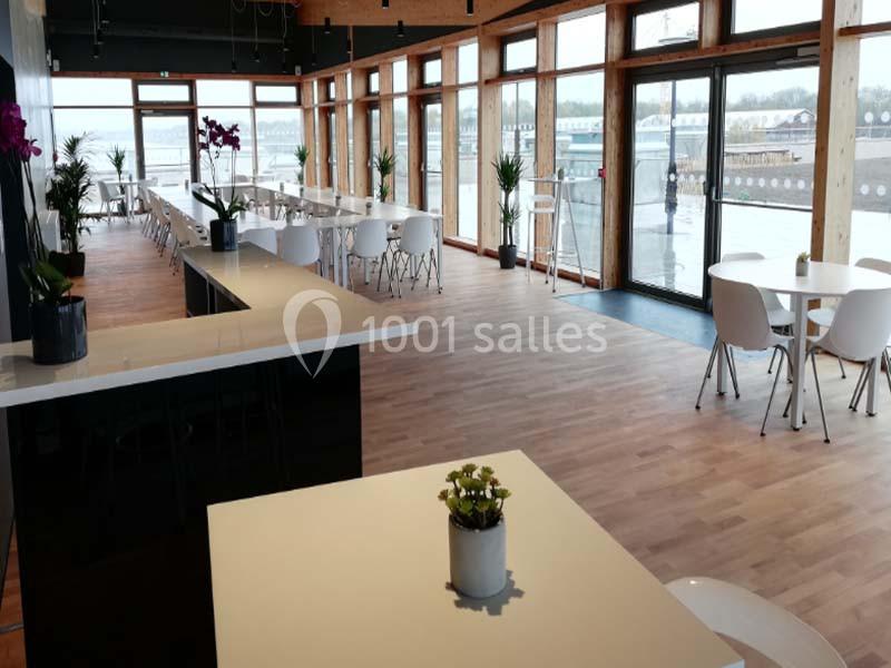 Salle lumineuse avec grandes baies vitrées, tables blanches, chaises modernes et plantes décoratives.