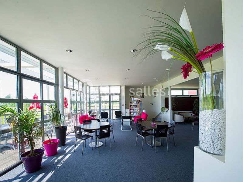 Salle lumineuse avec grandes baies vitrées, tables et chaises noires, plantes décoratives et étagère au fond.