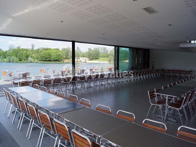 Salle spacieuse avec de grandes baies vitrées donnant sur un paysage de lac et des tables alignées avec des chaises en bois.