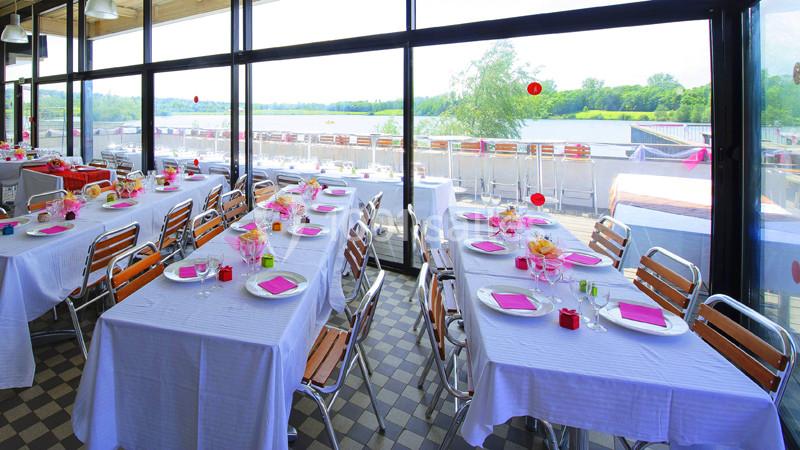 Salle de réception lumineuse avec grandes baies vitrées, tables dressées avec nappes blanches et vue sur un lac.