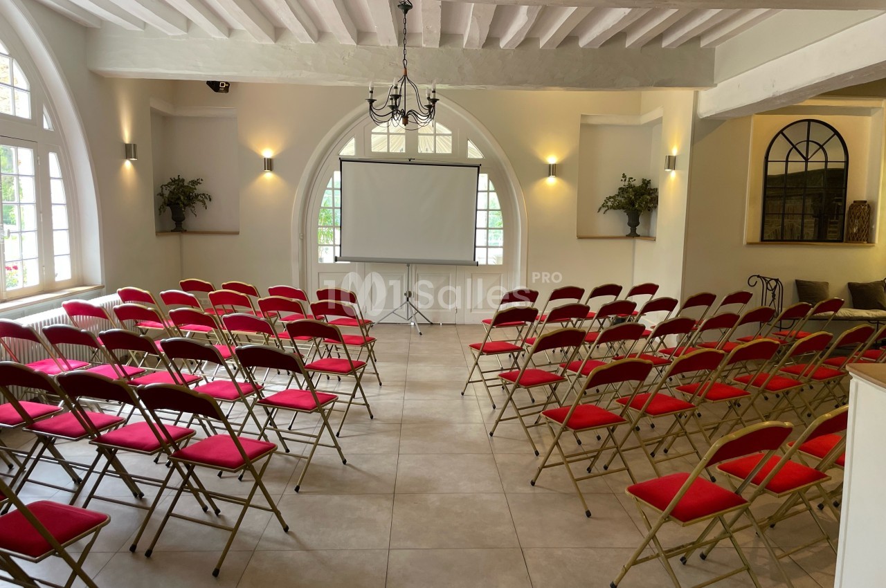 Salle lumineuse avec chaises rouges alignées face à un écran de projection, décorée de plantes et de grandes fenêtres.
