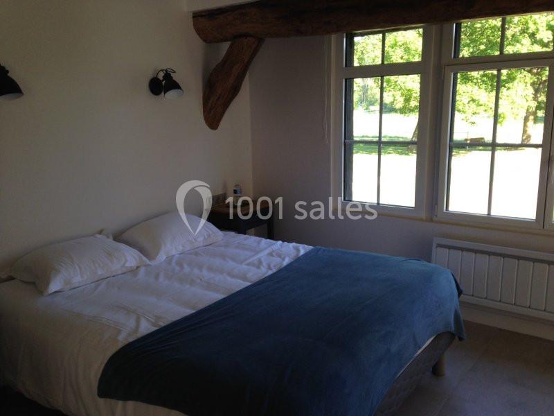 Chambre lumineuse avec un lit double, une couverture bleue, des murs clairs et une grande fenêtre donnant sur un espace vert.