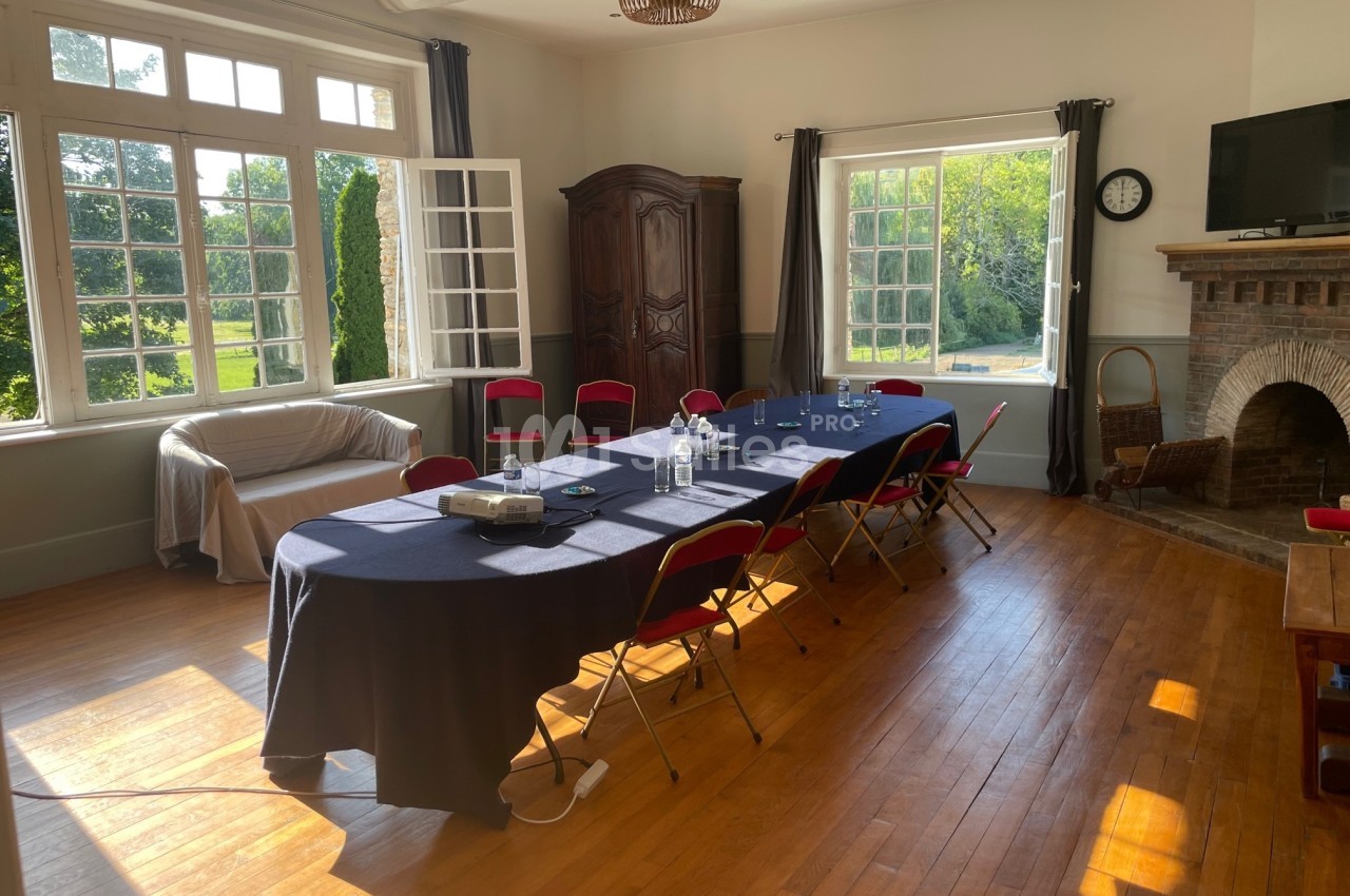 Salle lumineuse avec table de réunion, chaises rouges, projecteur, cheminée, armoire et grandes fenêtres donnant sur un…