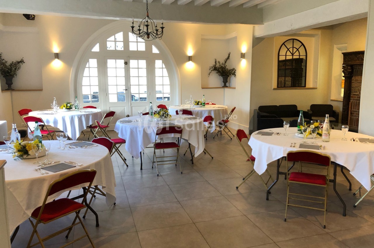 Salle lumineuse avec tables rondes dressées, chaises rouges, grandes fenêtres et décoration sobre.