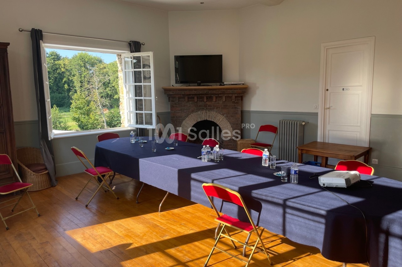 Salle lumineuse avec une grande table rectangulaire couverte d'une nappe bleue, entourée de chaises rouges, et une fenêtre…