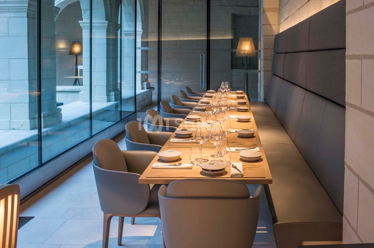 Salle de restaurant moderne avec une longue table dressée, chaises grises et grandes baies vitrées donnant sur une cour.