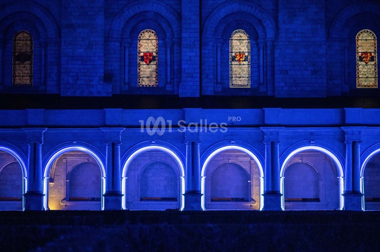 Façade d'un bâtiment en pierre avec des arches illuminées en bleu et des vitraux colorés visibles en hauteur.