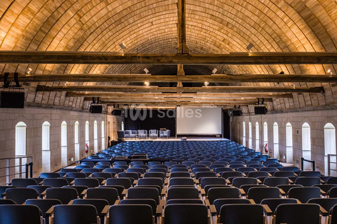 Salle de conférence avec rangées de sièges vides, écran de projection et plafond voûté en bois.