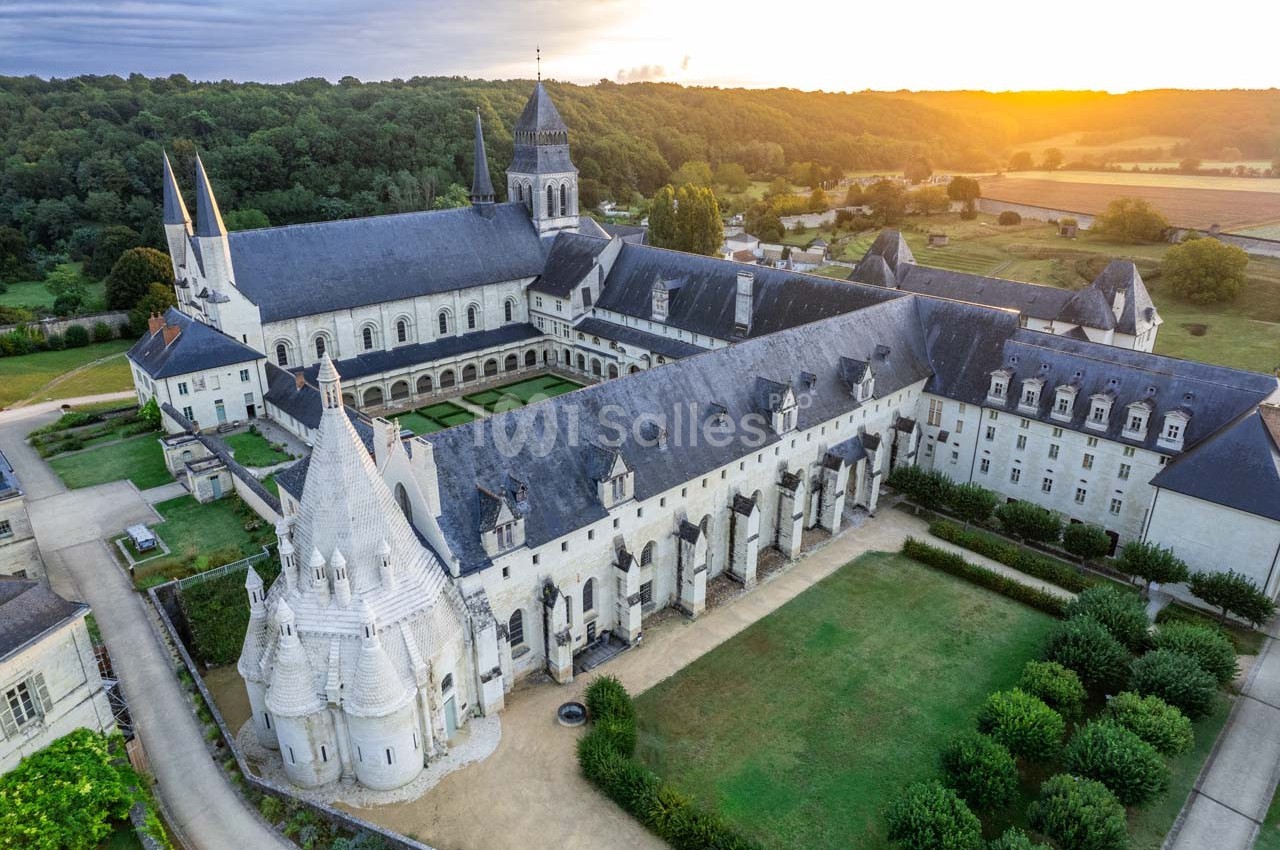 Vue aérienne d'une abbaye médiévale entourée de jardins et de champs, éclairée par une lumière de coucher de soleil.