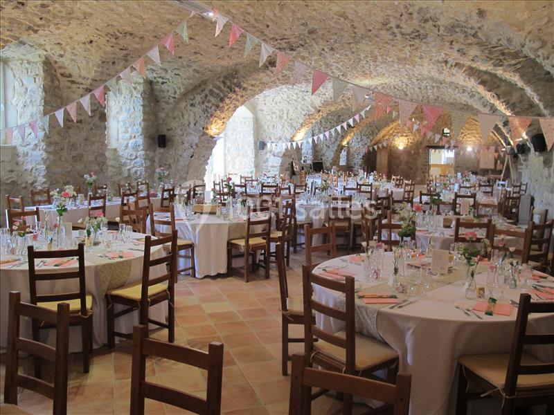 Salle voûtée en pierre décorée pour un événement, avec des tables rondes dressées et des guirlandes suspendues.