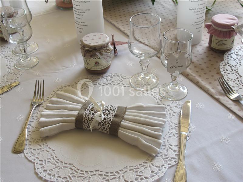 Table dressée avec nappe blanche, serviette pliée avec un ruban décoratif, couverts, verres et pots décoratifs.