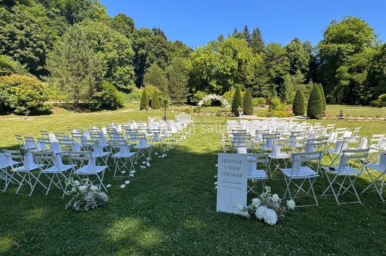 Chaises blanches disposées en extérieur pour une cérémonie, entourées de verdure et décorées de fleurs blanches.