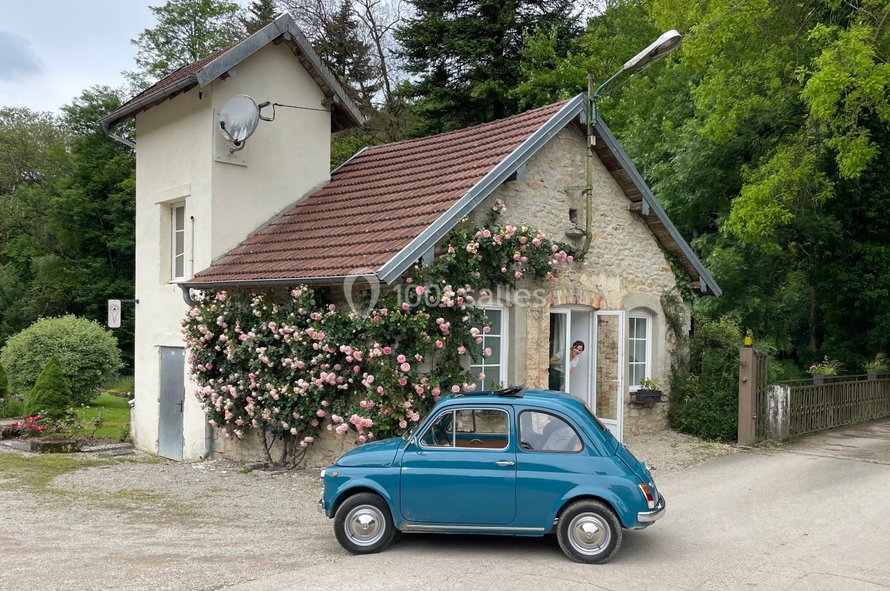 Petite voiture bleue garée devant une maison en pierre ornée de rosiers fleuris, entourée de verdure.