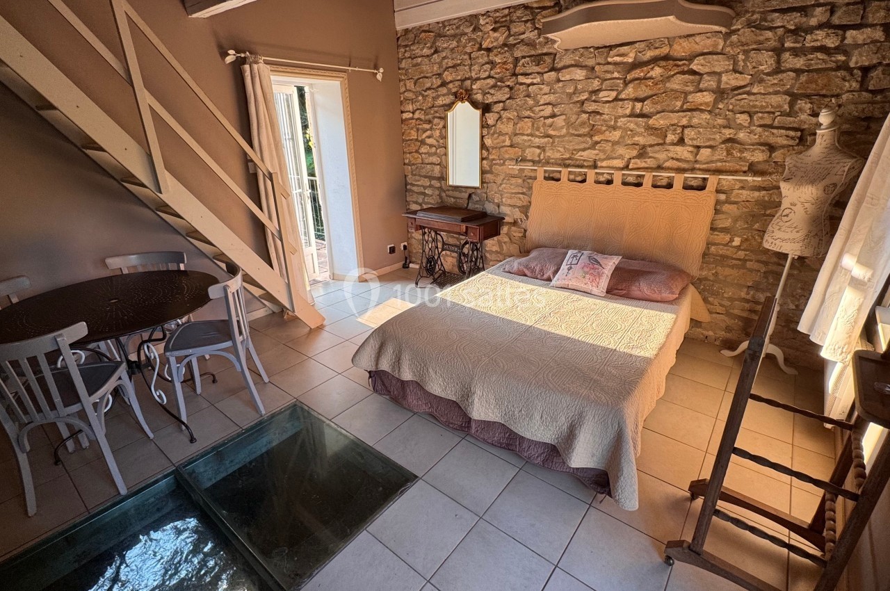 Chambre lumineuse avec lit double, mur en pierre, table ronde, escalier en bois et mannequin décoratif.