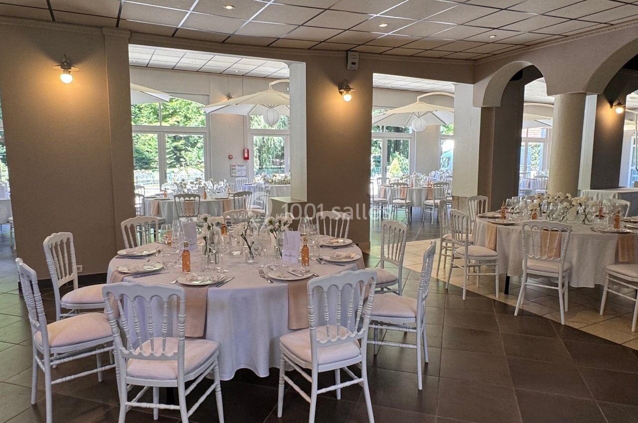 Salle de réception lumineuse avec tables rondes dressées, nappes blanches et chaises élégantes, vue sur l'extérieur…
