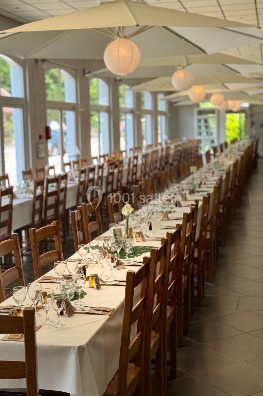 Salle de réception lumineuse avec de longues tables dressées, nappes blanches et chaises en bois alignées.