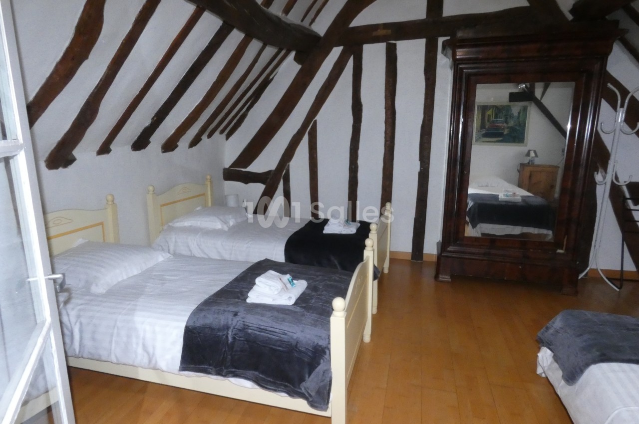 Chambre avec poutres apparentes, deux lits simples, sol en bois, armoire avec miroir et lumière naturelle.