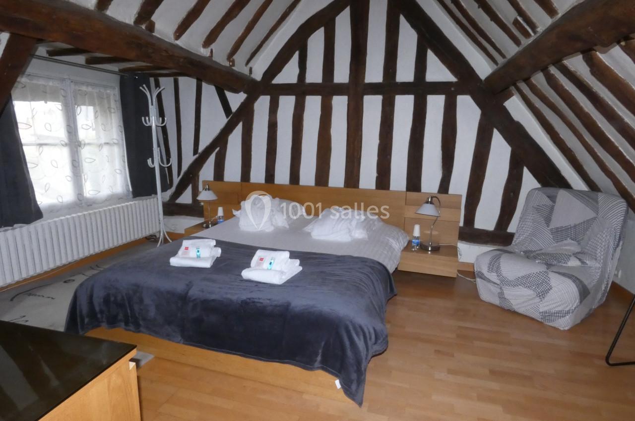 Chambre avec poutres apparentes, lit double, fauteuil, parquet et lumière naturelle provenant d'une fenêtre.