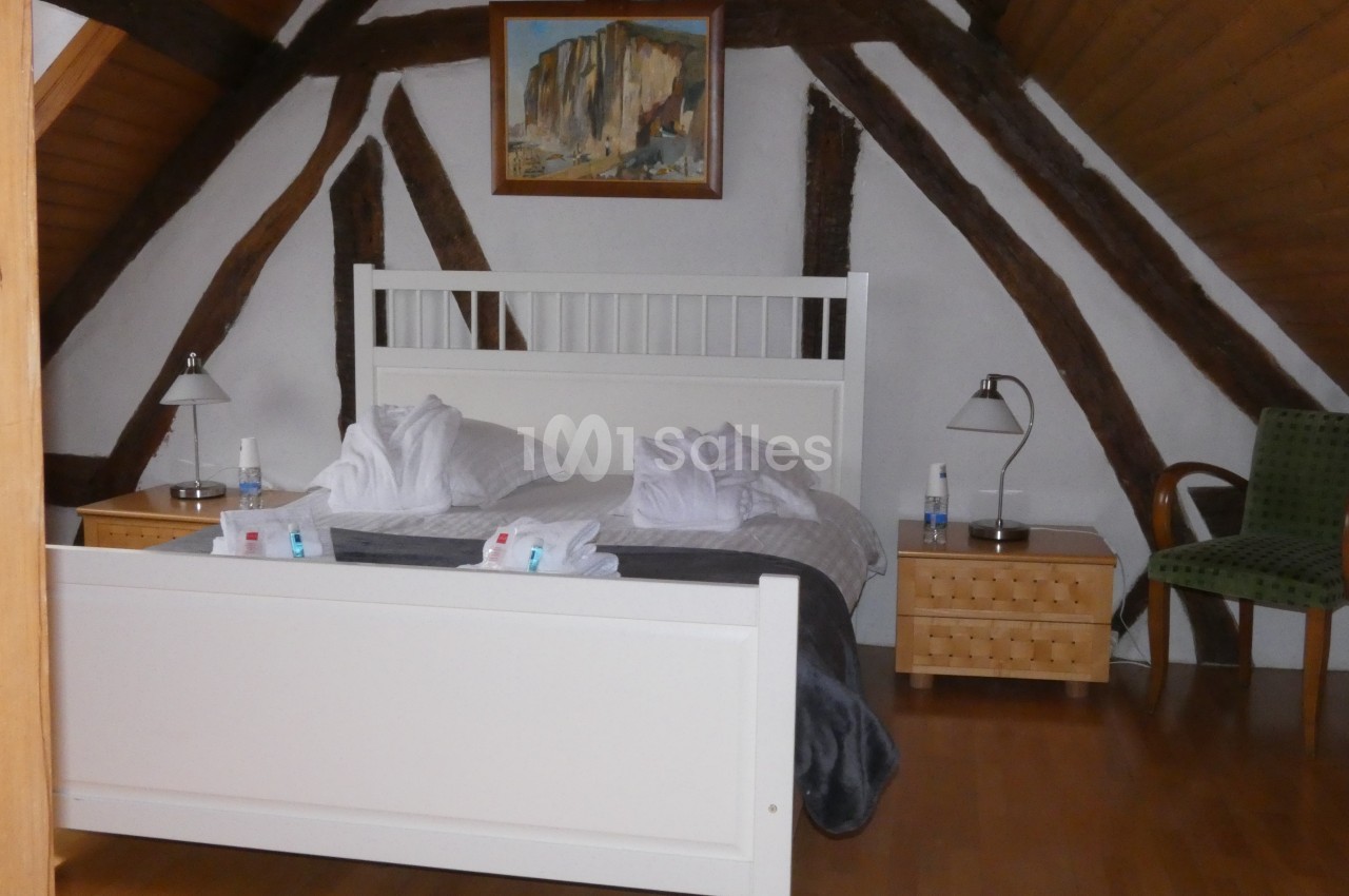 Chambre avec poutres apparentes, lit double, tables de chevet, lampes, fauteuil et tableau au mur.