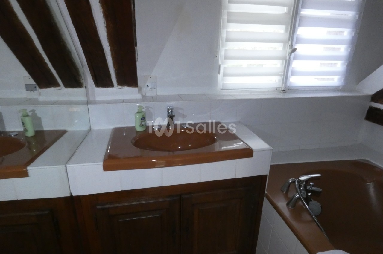 Salle de bain avec lavabo marron, miroir, baignoire équipée d'une douchette et fenêtre à persiennes blanches.