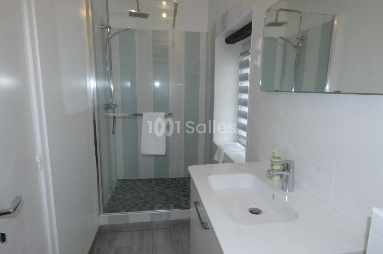 Salle de bain moderne avec douche à l'italienne, lavabo blanc et miroir, éclairée par une fenêtre avec store.