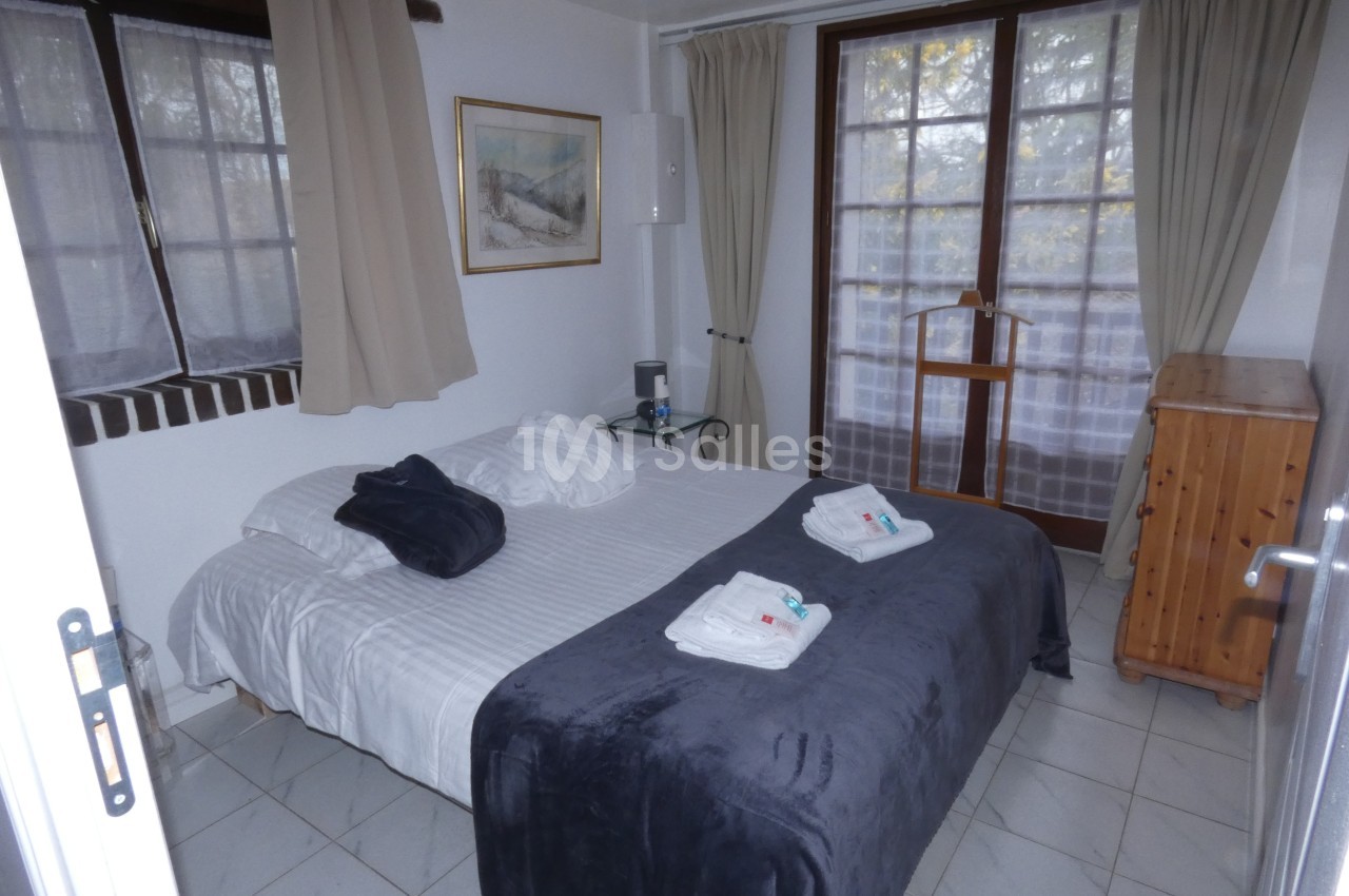 Chambre avec lit double, couverture grise, fenêtres avec rideaux clairs et mobilier en bois.