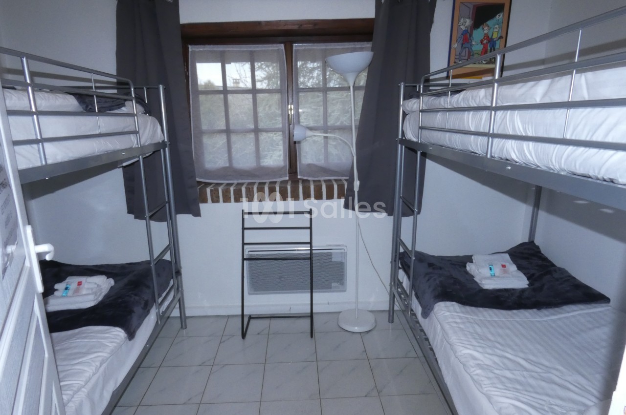 Chambre avec deux lits superposés, fenêtre centrale, rideaux gris, sol carrelé et lampadaire blanc.