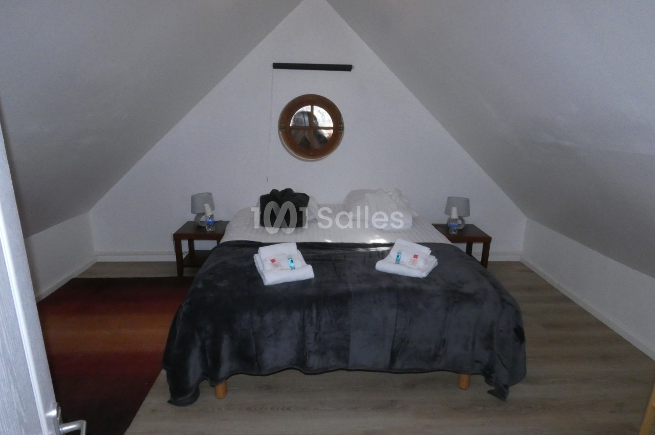 Chambre mansardée avec un lit double, draps pliés, tables de chevet avec lampes et un miroir rond au mur.