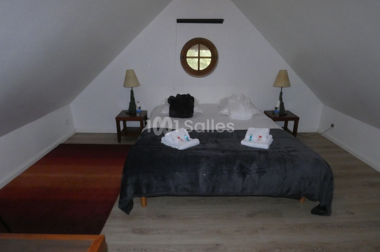 Chambre mansardée avec lit double, deux lampes de chevet, fenêtre ronde et sol en parquet clair.