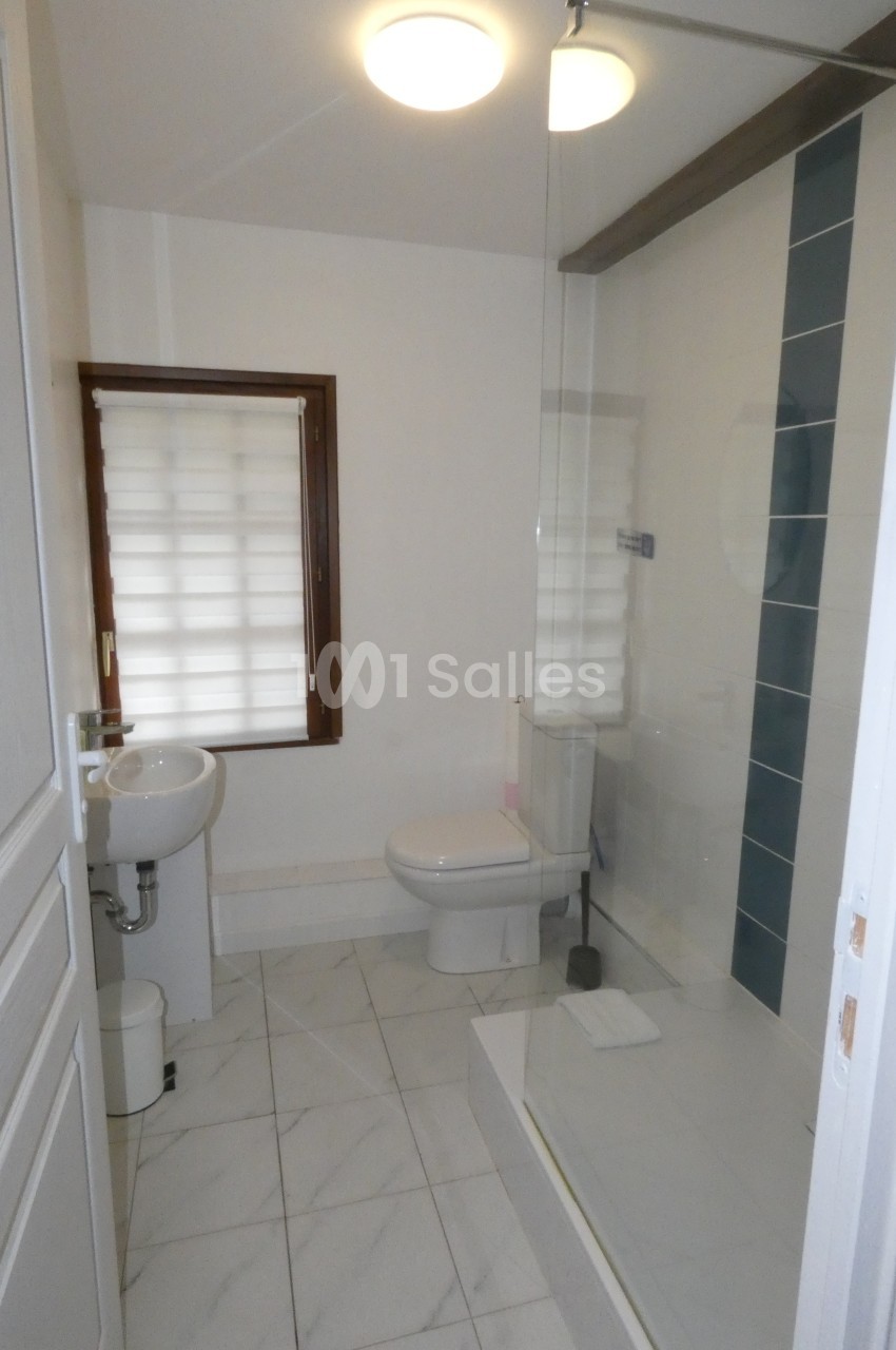 Salle de bain avec douche à l'italienne, lavabo, toilettes, carrelage blanc et fenêtre avec store.