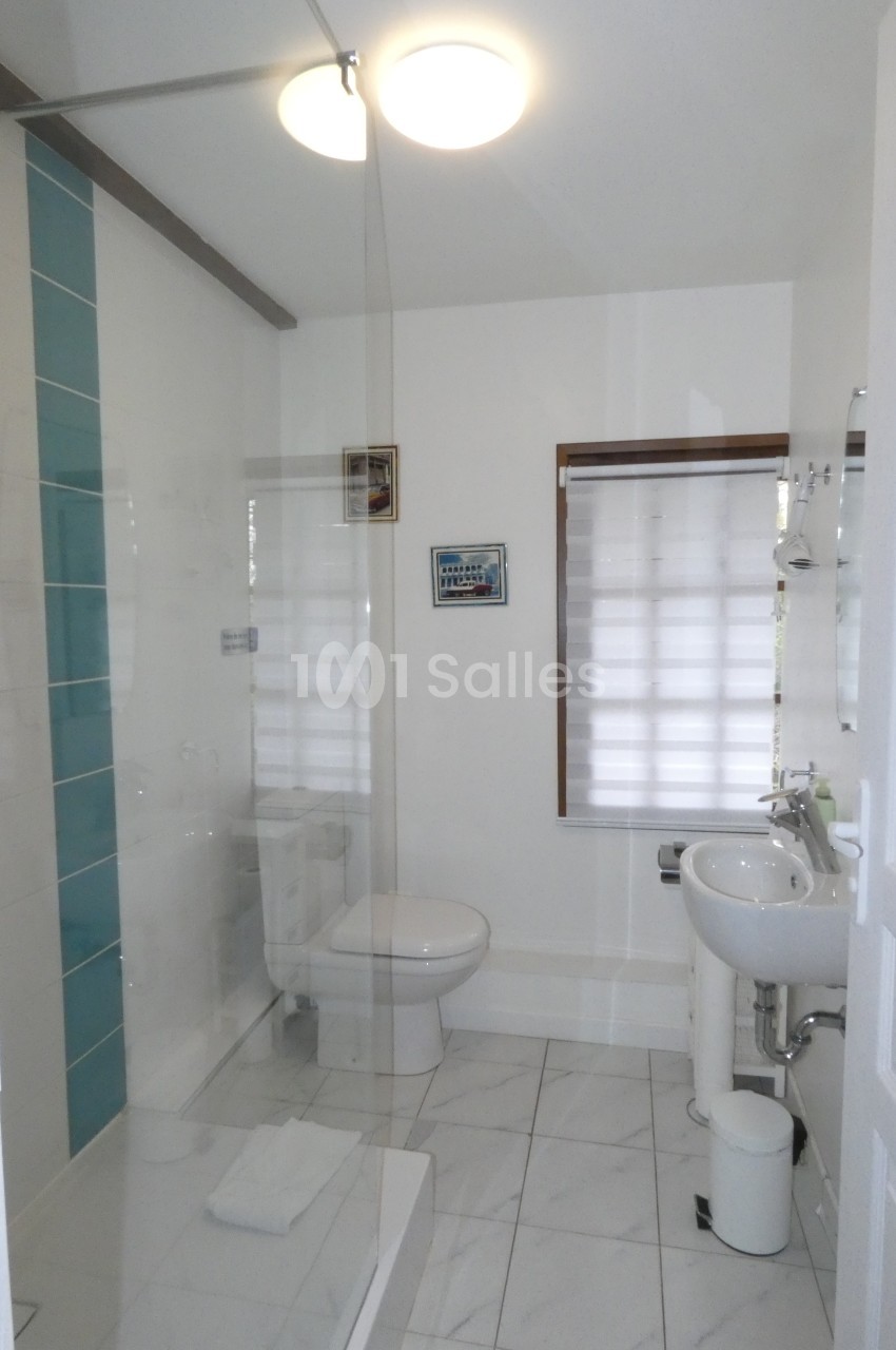 Salle de bain lumineuse avec douche à l'italienne, lavabo, toilettes et fenêtre avec store semi-ouvert.
