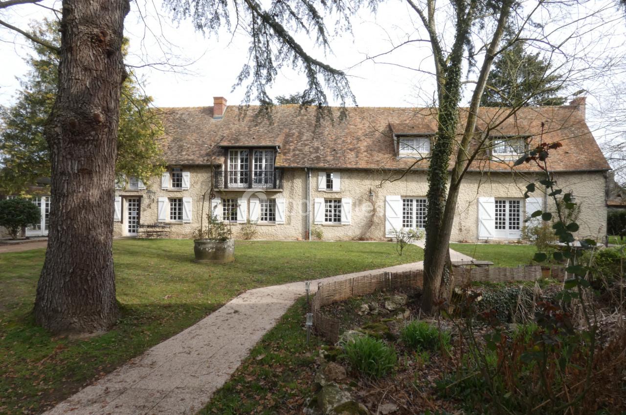 Grande maison en pierre avec toit en tuiles, entourée d'un jardin arboré et d'un chemin en gravier.