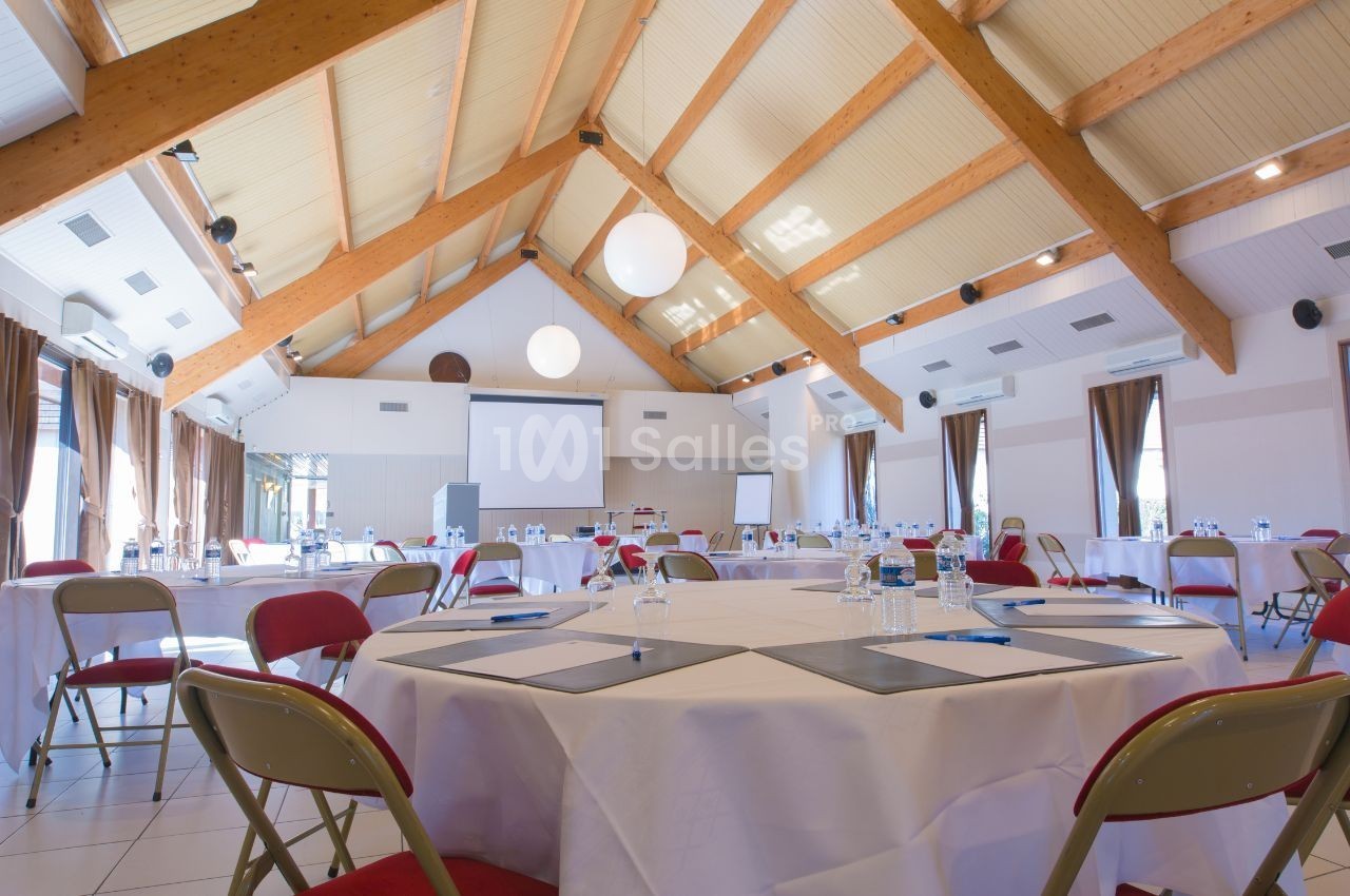 Salle de réunion lumineuse avec tables rondes dressées, chaises pliantes et projecteur installé devant un écran blanc.