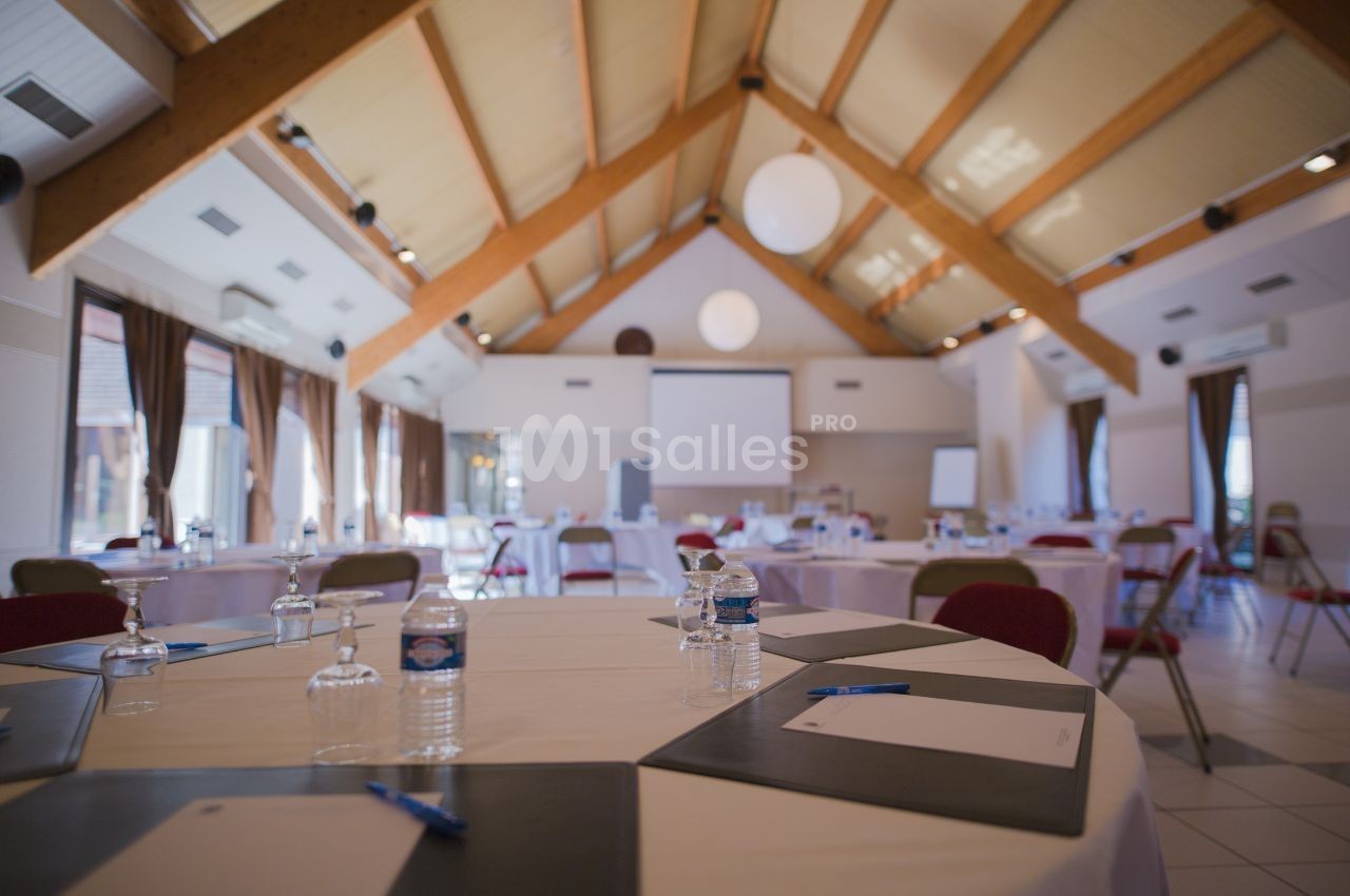 Salle de réunion lumineuse avec tables rondes dressées, chaises, écran de projection et grandes fenêtres.