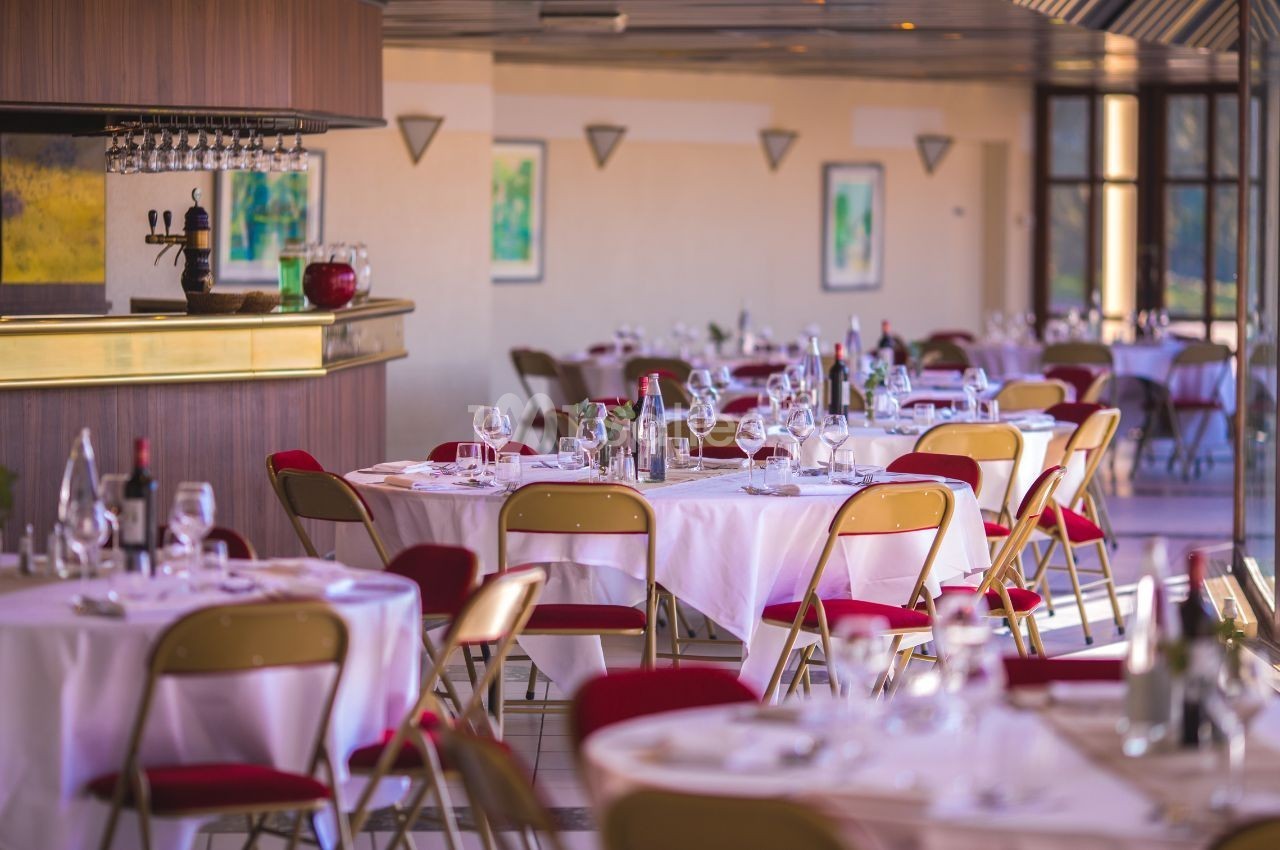 Salle de restaurant lumineuse avec tables dressées, nappes blanches, chaises rouges et un bar en arrière-plan.
