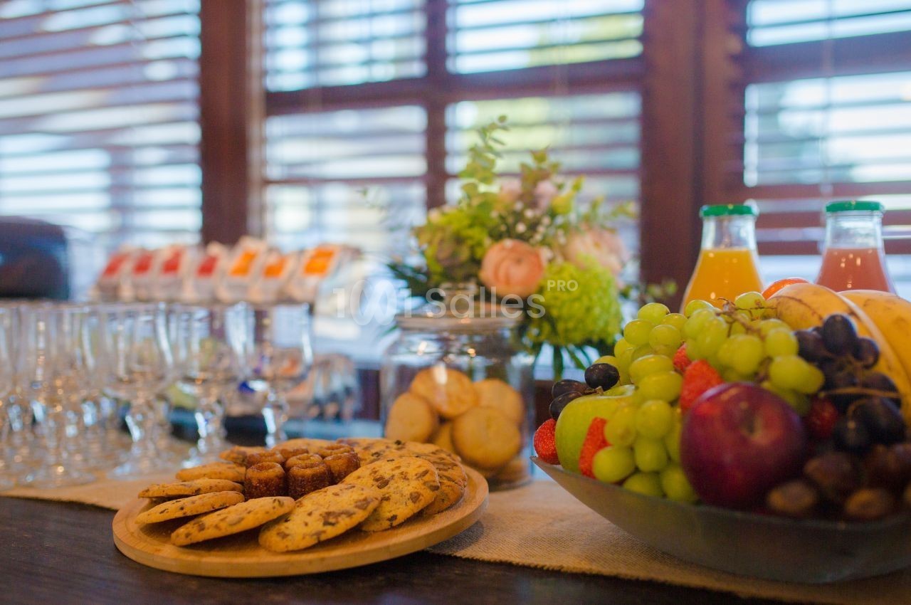 Plateau de biscuits et fruits frais sur une table, avec des boissons et des verres en arrière-plan dans une salle lumineuse.