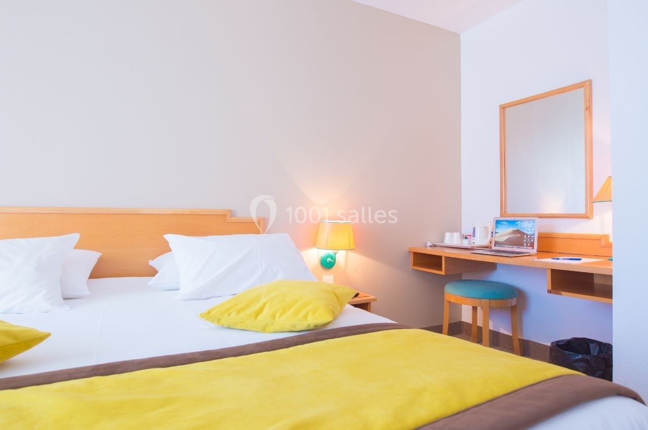 Chambre lumineuse avec lit double, coussins jaunes, bureau en bois, miroir et lampe de chevet allumée.