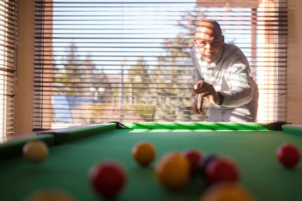 Un homme joue au billard dans une pièce lumineuse avec des stores laissant passer la lumière extérieure.