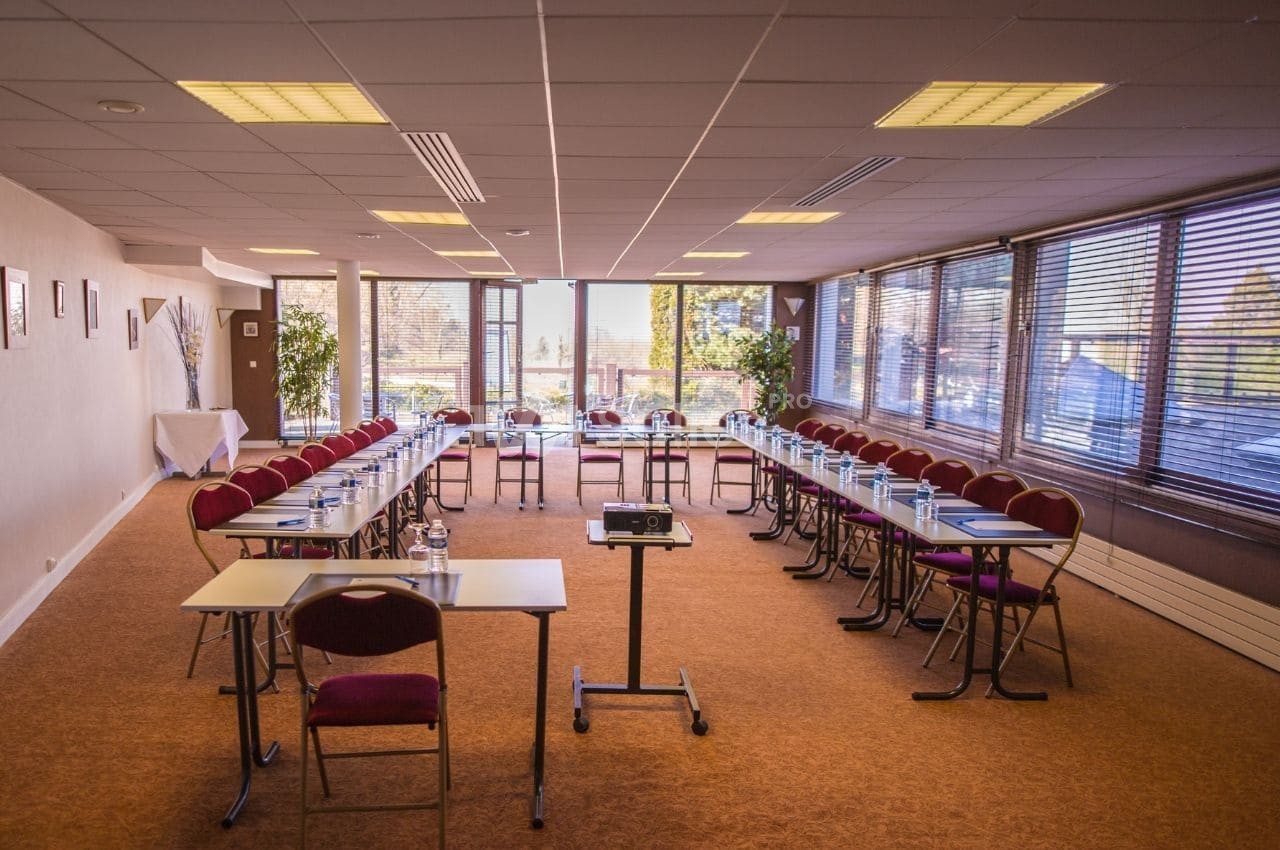 Salle de réunion lumineuse avec tables disposées en U, chaises rouges, projecteur et vue sur l'extérieur.