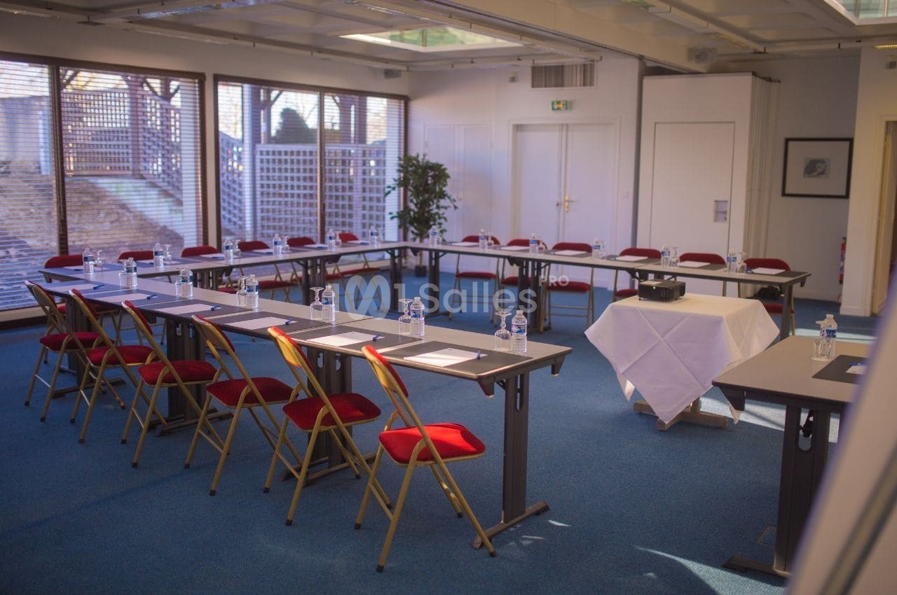 Salle de réunion lumineuse avec tables disposées en U, chaises rouges et matériel de conférence sur les tables.