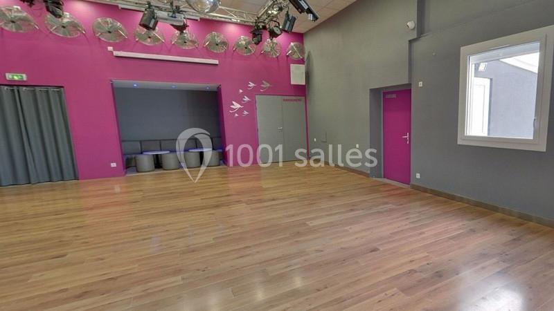 Salle lumineuse avec parquet, murs gris et roses, chaises alignées dans un coin, éclairage suspendu au plafond.