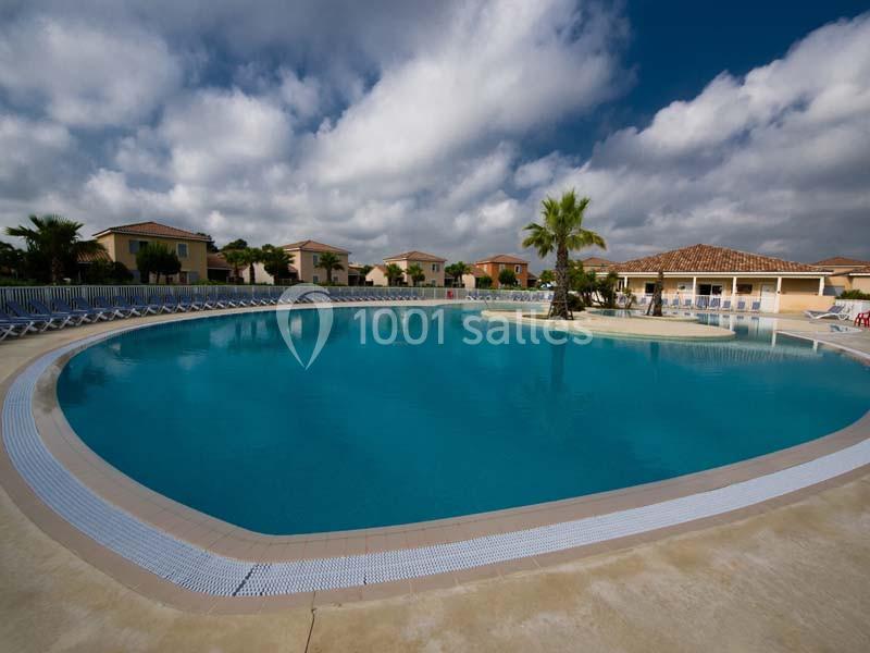 Piscine extérieure circulaire entourée de transats, avec des maisons et des palmiers en arrière-plan sous un ciel nuageux.