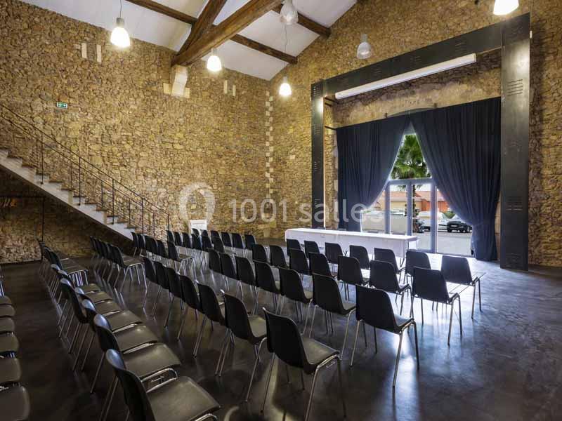 Salle de conférence en pierre avec chaises noires alignées, une scène et des rideaux ouverts sur une vue extérieure.