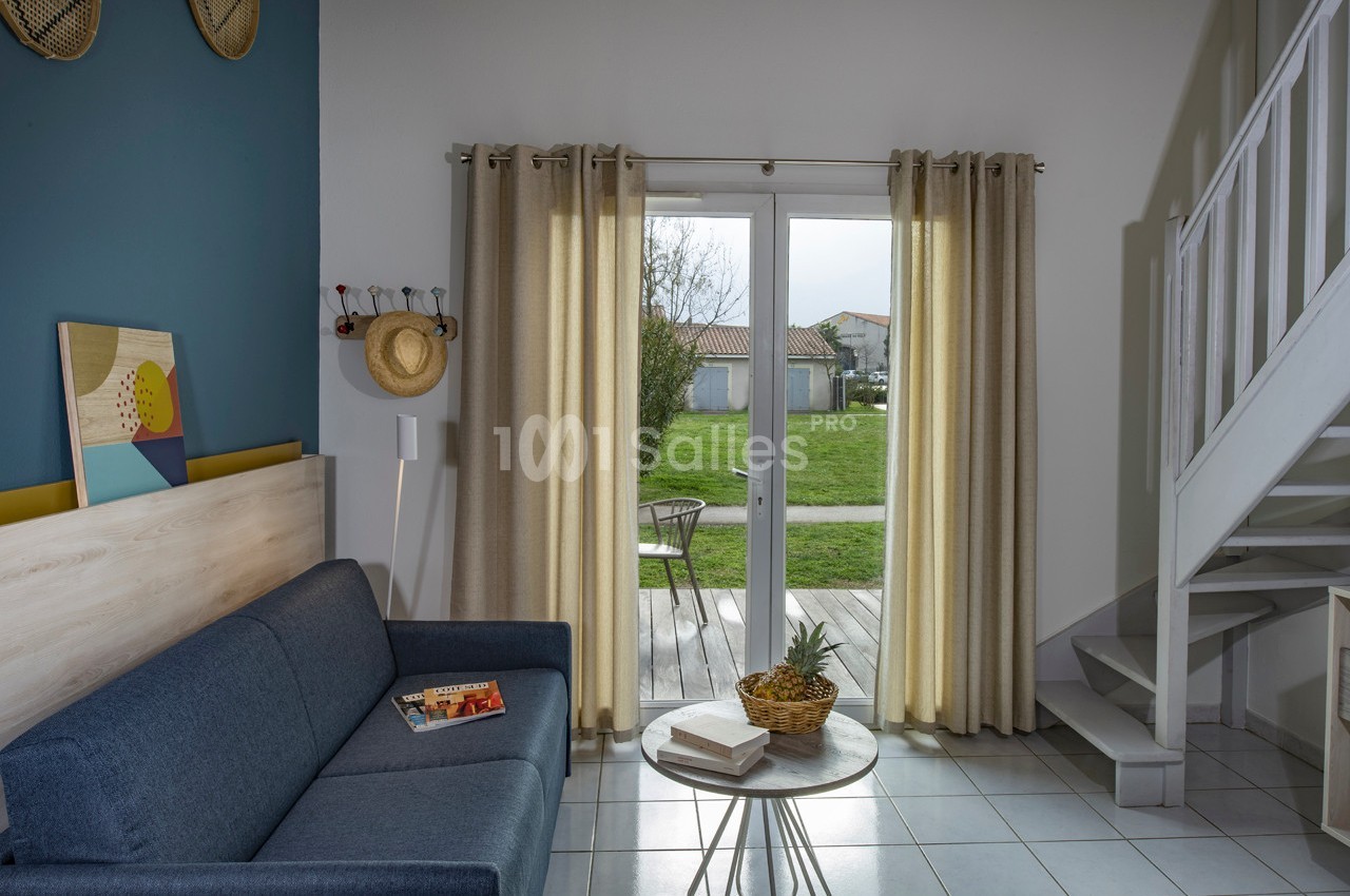 Salon lumineux avec canapé bleu, table basse, porte-fenêtre donnant sur une terrasse et un jardin verdoyant.