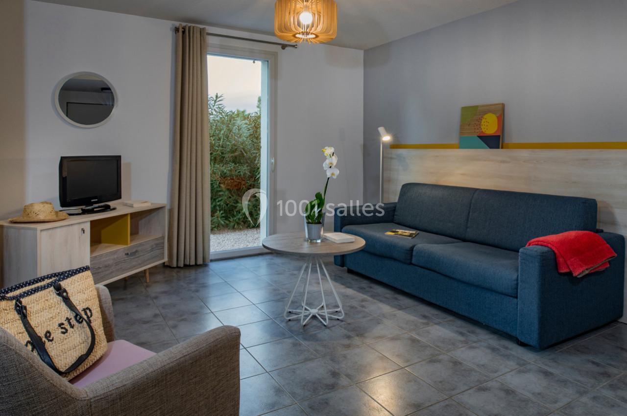 Salon lumineux avec canapé bleu, table basse, fauteuil, télévision, et porte-fenêtre donnant sur un jardin.