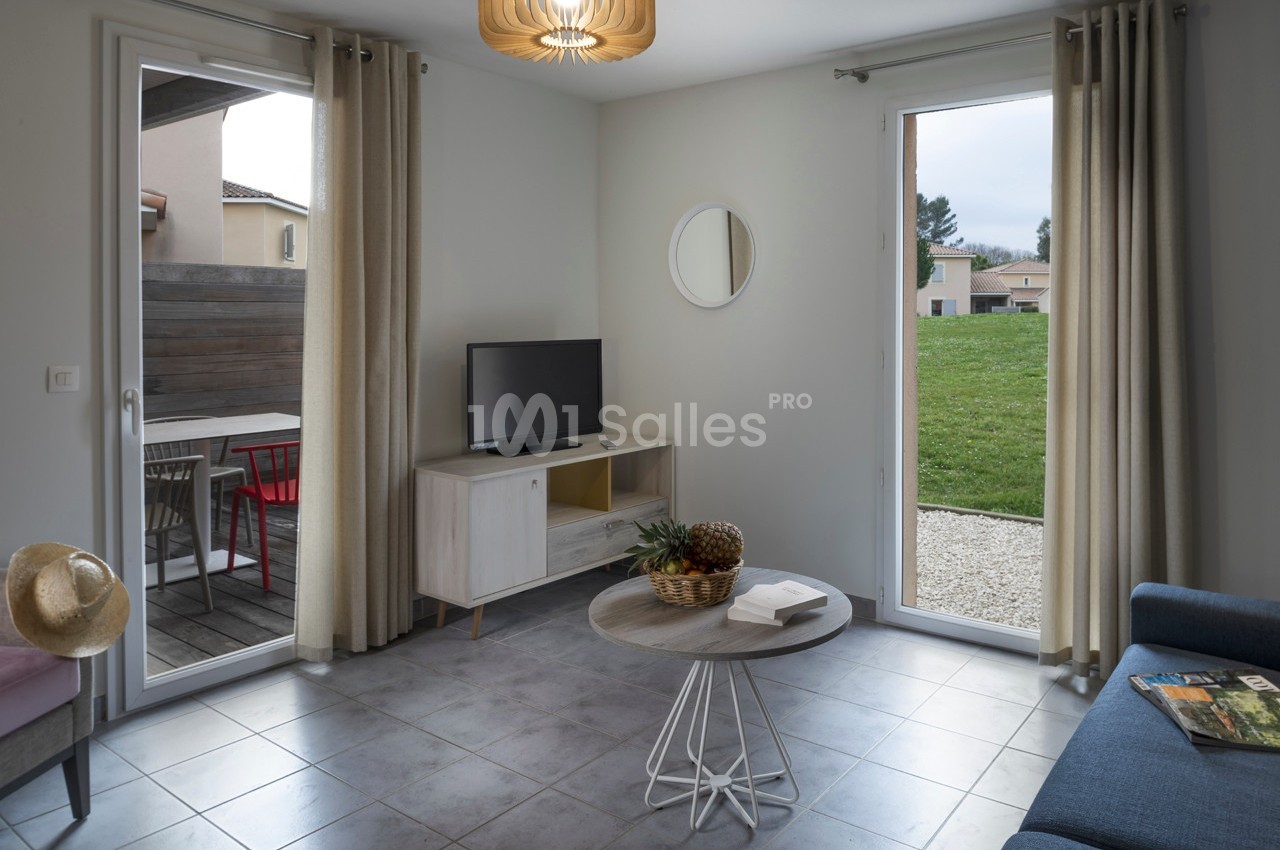 Salon lumineux avec canapé, table basse, télévision, grandes baies vitrées donnant sur une terrasse et un jardin.